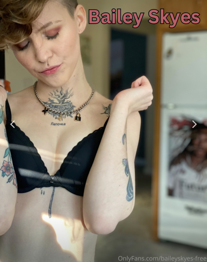 Bailey Skyes is a tattooed model. She is wearing a black bra and pushing her small breasts together while looking down at them.